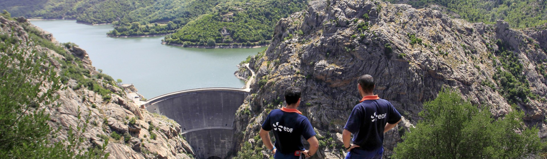 Vue sur le barrage de Tolla en Corse, avec deux agents EDF en tenue de travail observant l’ouvrage hydraulique et le lac entouré de reliefs montagneux.