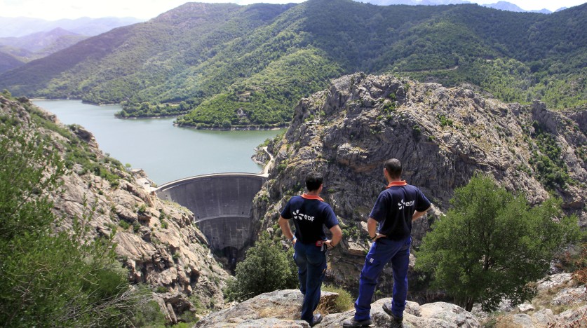 Vue sur le barrage de Tolla en Corse, avec deux agents EDF en tenue de travail observant l’ouvrage hydraulique et le lac entouré de reliefs montagneux.